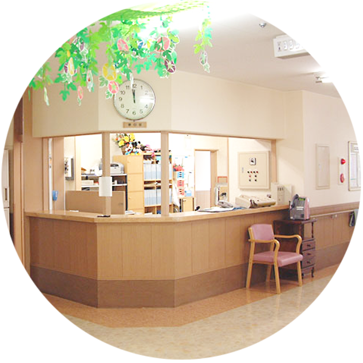 木製カウンター受付と待合スペース。時計と装飾植物が設置された明るい介護施設のロビー。
